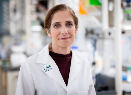 Dr. Casey Cook wearing a black shirt with a white lab coat standing in front of a laboratory setup that is blurry. 