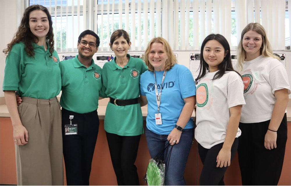 Group of six people standing indoors, some wearing green Adopt a School shirts, others in blue AVID and white Activist Lab shirts.