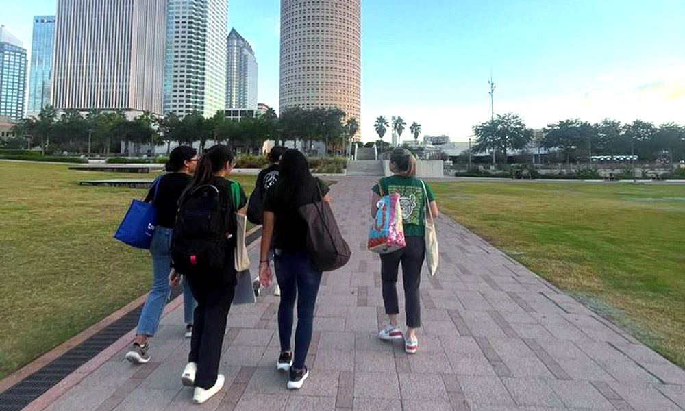 A group of women walking on a pathed walkway towards buildings downtown. 