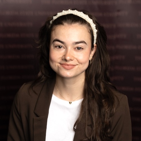 A woman with long brown hair smiles wearing a brown jacket and white shirt