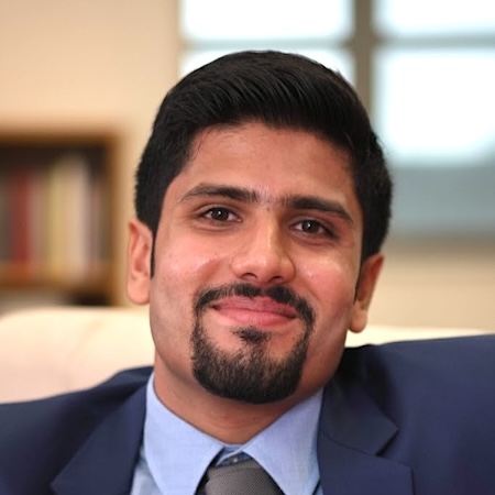 A man with dark hair and a goatee smiles in a suite